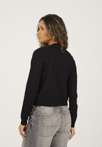 Vrouw met krullend haar, gekleed in een zwart shirt met lange mouwen en lichtgrijze jeans, staand en kijkend naar links tegen een effen achtergrond.