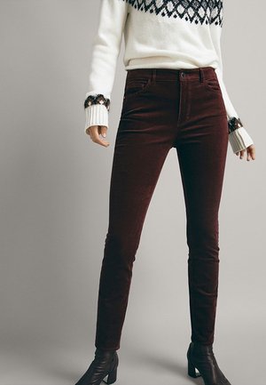 Femme portant un pantalon en velours côtelé marron foncé, un pull en maille blanche avec un motif noir aux poignets et aux épaules, et des bottines noires à talons.