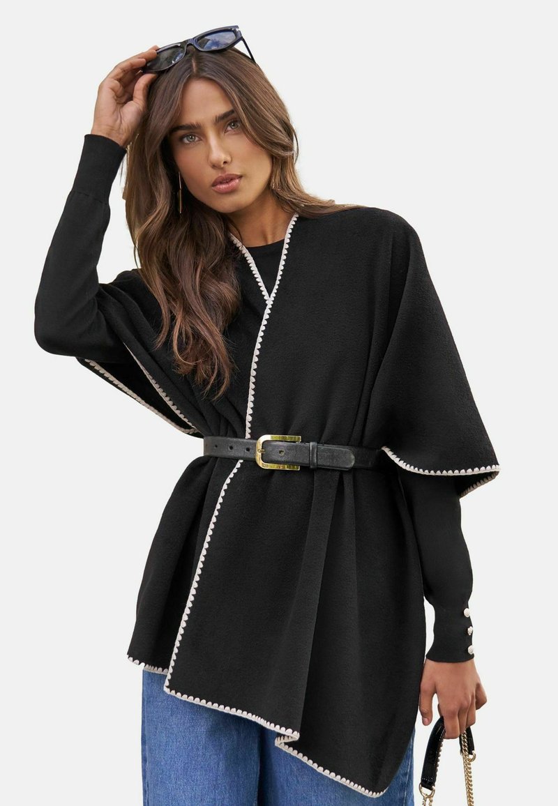 Pelerine neagră în stil poncho cu trim alb, cu o talie strânsă cu o curea neagră, mâneci lungi și o textură moale.