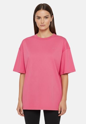 Donna con capelli castani lisci che indossa una t-shirt rosa a maniche corte e pantaloni neri, in piedi davanti a uno sfondo bianco semplice.