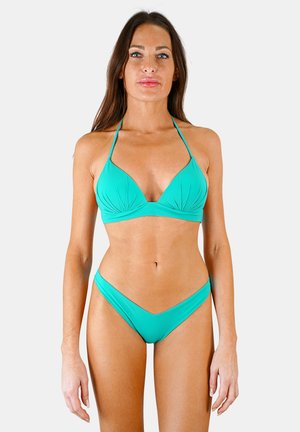 Femme debout, face à l'objectif, portant un bikini deux pièces turquoise sur un fond blanc uni.