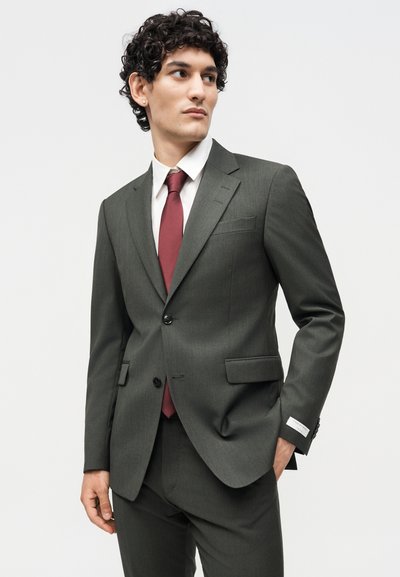 Νέος άνδρας φοράει σκούρο γκρι κοστούμι, λευκό πουκάμισο και μπορντό γραβάτα, στέκεται με το ένα χέρι στην τσέπη, κοιτάζοντας προς τα πλάγια σε απλό φόντο.