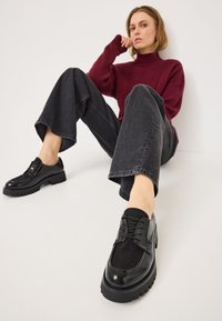 Schwarze Schnürschuhe mit glänzender Oberfläche und Wildlederakzenten, ausgestattet mit einer dicken, strukturierten Sohle. Das Design ist rundlich mit einer geschmeidigen Silhouette.
