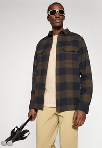 Man in een oversized geruite jas en beige broek, met zwarte wandelstokken in de hand, en grote beige zonnebril op een neutrale achtergrond.