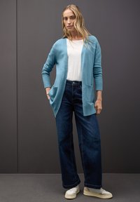 Ljusblå stickad cardigan med ribbade manschetter och fickor, matchad med mörkblå vida jeans och vita sneakers. Slät textur, avslappnad passform.