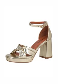 Sandalias de tacón metálicas doradas con una amplia tira que presenta un diseño anudado. Tacón grueso y correa al tobillo con hebilla ajustable. Textura suave.