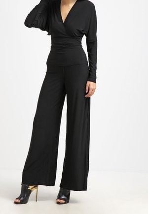 Schwarzer Overall mit langen Ärmeln, Wickeldesign an der Taille, weit geschnittene Silhouette, aus weichem Stoff; kombiniert mit schwarzen, offenen Absätzen.