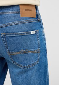 Blå denimjeans med en brun läderbaksplatta märkt "MUSTANG." Funktioner inkluderar subtil sömnad och en bakficka med berg-formad broderi.