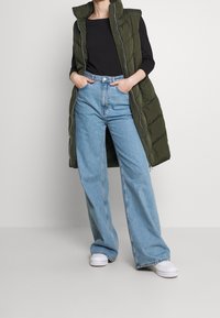 Oliwkowo zielona pikowana kamizelka bez rękawów na czarnej koszulce, zestawiona z jasno-niebieskimi dżinsami o szerokich nogawkach. Całość uzupełniają białe trampki.