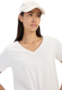 Biała koszulka V-neck z miękkiego, lekkiego materiału, połączona z kremową czapką z naszytym okrągłym logo na przodzie.