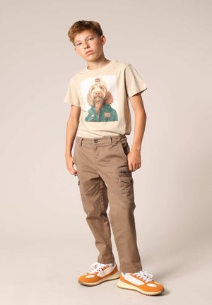 Beige T-shirt met een hondenafbeelding, gecombineerd met bruine cargobroeken. Sneakers met oranje accenten en een chunky zool. Eenvoudig, functioneel design.