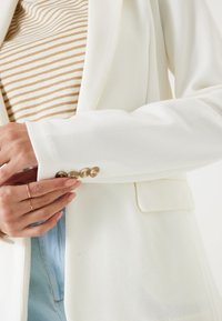 Biała marynarka z teksturowanej tkaniny, jednorzędowa, z złoconymi guzikami na mankietach; zestawiona z paskowanym, beżowym topem.