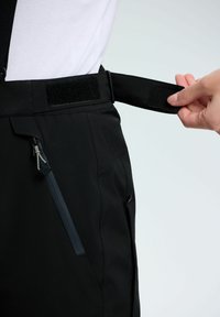 Eine Hand passt den verstellbaren Bund einer schwarzen Hose mit Klettverschluss an, wobei die seitliche Reißverschlusstasche sowie ein Teil eines weißen Shirts und Hosenträger zu sehen sind.