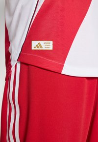 Röda sportshorts med vita detaljer, med ett andningsbart nätpanel, tre vita ränder på sidan och en Adidas äkthetsetikett.