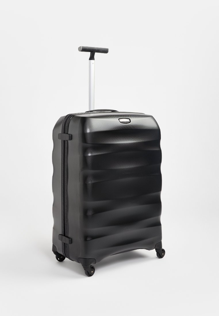 Valise rigide noire avec un design à vagues texturé, une poignée argentée rétractable, des doubles roulettes et une fermeture éclair supérieure.