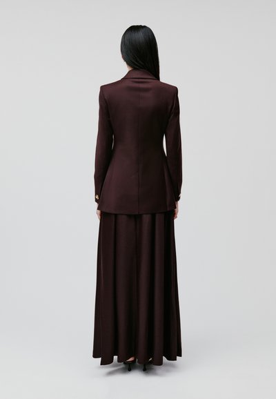 Długa burgundowa kurtka z wyrazistymi klapami na tle pasującej, długiej spódnicy. Gładka tekstura, dopasowany krój i przyciski na rękawach.