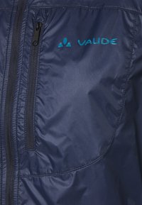 Mörkblå Vaude-jacka med en slät, glänsande yta. Har en frontdragkedja och synlig logotyp i ljusblått. Utformad för utomhusbruk.