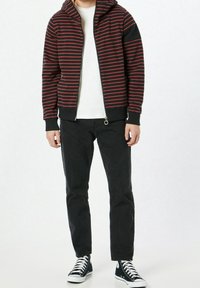 Sudadera con capucha de rayas rojas y negras, cerrada con cremallera, usada sobre una camiseta blanca. Combinada con pantalones negros y zapatillas altas negras.