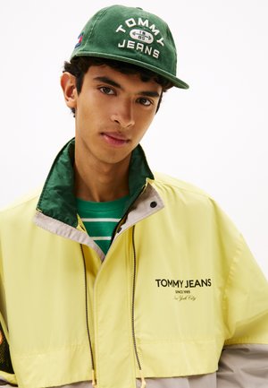 Geel en beige windjack met groene kraag en "TOMMY JEANS" logo. Groene pet met witte tekst. Daaronder een groen gestreept shirt.