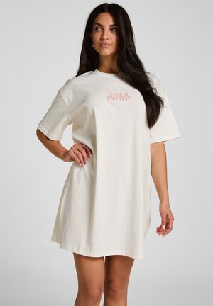Vrouw met lang donker haar die een losse, beige jurk met korte mouwen draagt met roze tekst op de borst waarop staat "SLEEP IN STAY COZY".
