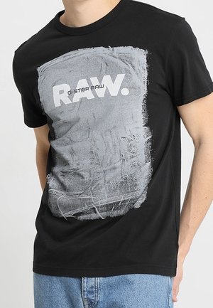 Czarna koszulka z białą i szarą abstrakcyjną grafiką oraz wyraźnym napisem "RAW." i mniejszym napisem "G-STAR RAW" z przodu, noszona przez osobę.