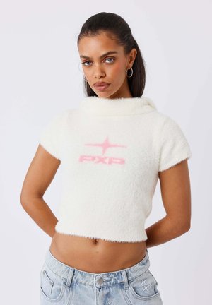 Femme aux cheveux foncés portant un top court blanc duveteux avec un logo rose et un jean denim bleu clair sur un fond uni.