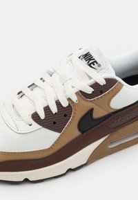 Nike Air Max tenisky v bílé, hnědé a béžové barvě s černým logem swoosh, bílými tkaničkami a viditelnou detailní vzduchovou tlumicí podrážkou.
