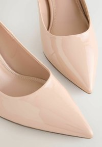 Naaktkleurige stilettohakken van lakleer met een puntige neus, voorzien van een gladde afwerking en een laag uitgesneden ontwerp aan de zijkanten.