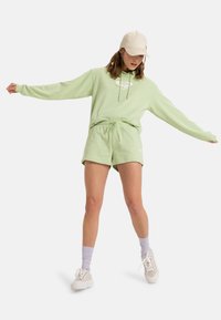 Sudadera verde claro con logo, pantalones cortos verdes a juego y zapatillas blancas. Tejido texturizado, corte relajado, acentuado con una gorra beige.