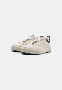 Light beige leather sneakers with textured rubber soles, navy accents, and beige laces. Smooth upper with rounded toe and low profile design.