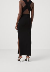 Femme portant une longue robe noire avec des bretelles croisées et une fente latérale, associée à des sandales à talons épais noires et argentées, debout sur un sol blanc.