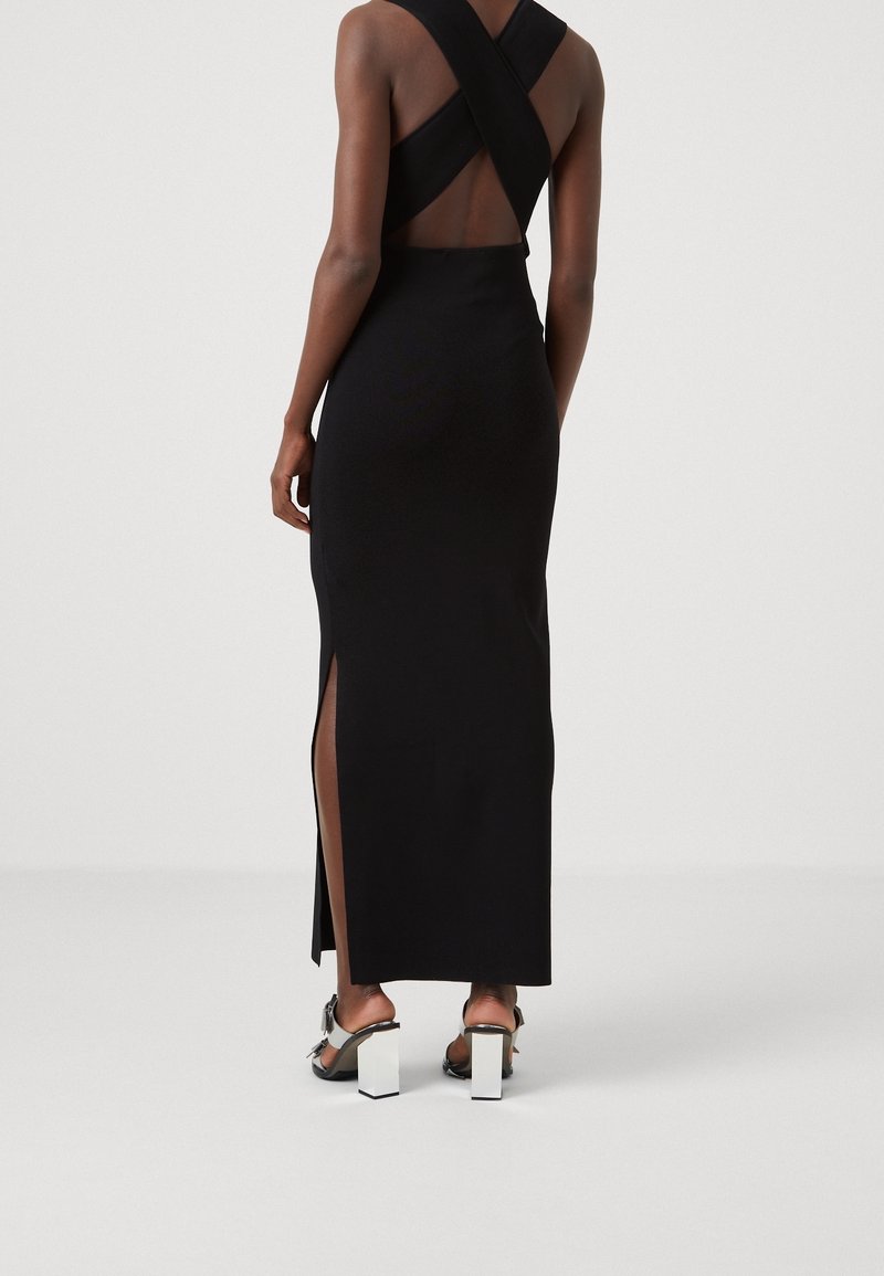 Femme portant une longue robe noire avec des bretelles croisées et une fente latérale, associée à des sandales à talons épais noires et argentées, debout sur un sol blanc.