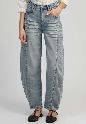 Femme portant un jean large bleu clair avec des détails délavés, une main dans la poche, associé à des sandales tissées noires et un haut blanc à manches en dentelle.