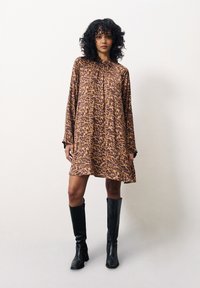 Tierdruckkleid in Braun und Beige, lockerer Schnitt mit langen Ärmeln, rundem Ausschnitt und leicht ausgestelltem Schnitt, kombiniert mit schwarzen knielangen Stiefeln.