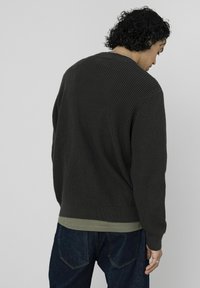 Pull en maille vert foncé avec un motif texturé, poignets et ourlet côtelés, superposé à une chemise vert clair, porté avec un jean bleu.