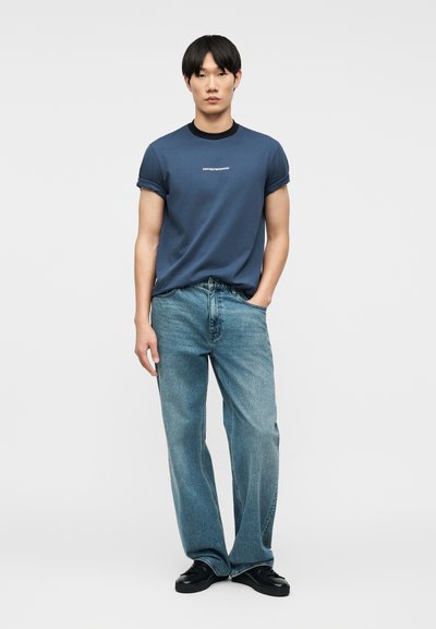 Νεαρός άνδρας που φοράει μπλε μπλουζάκι Emporio Armani με γυρισμένα μανίκια, φαρδιά ανοιχτό μπλε τζιν και μαύρα παπούτσια, στέκεται με το ένα χέρι στην τσέπη.