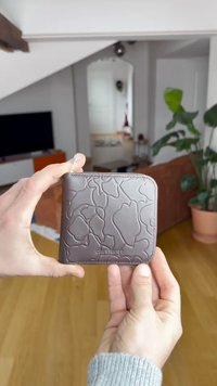 Brown rectangular wallet featuring a textured floral pattern, smooth material, and a zipper closure, held in a hand against a blurred background.