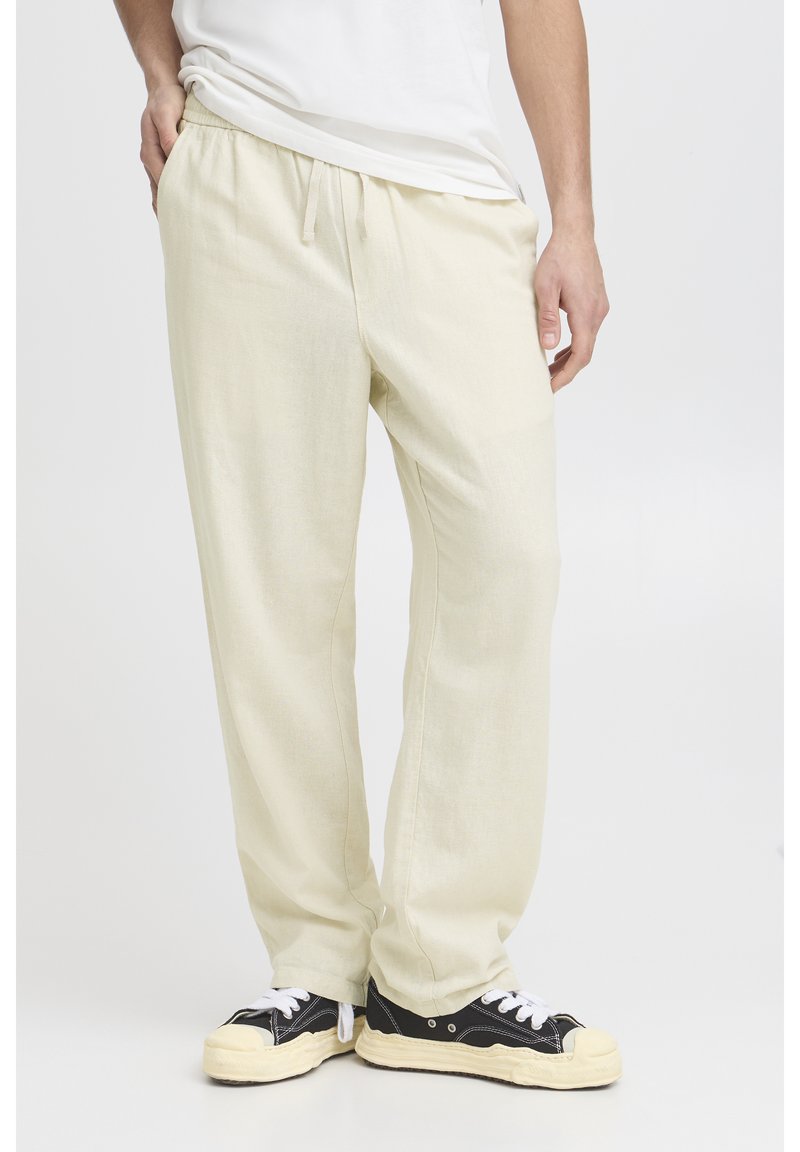 Light beige pants with an elastic waistband, drawstring detail, and a relaxed fit, paired with black and white sneakers.