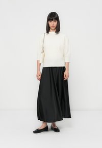 Femme se tenant debout, portant un pull blanc, une longue jupe noire, des ballerines noires et un sac à épaule blanc, devant un fond blanc uni.