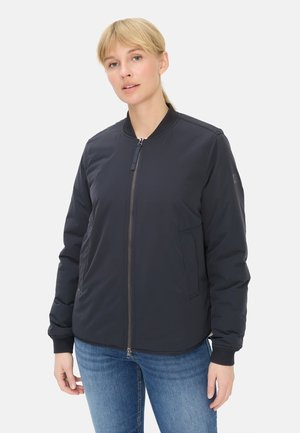 Femme blonde portant une veste bomber noire zippée avec des poignets côtelés et un jean bleu, debout devant un fond blanc uni.