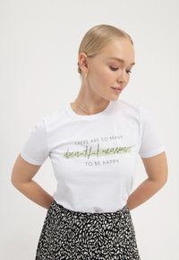 Hvid bomulds t-shirt med korte ærmer, med grøn og sort tekst, der lyder "Der er så mange smukke grunde til at være glad."