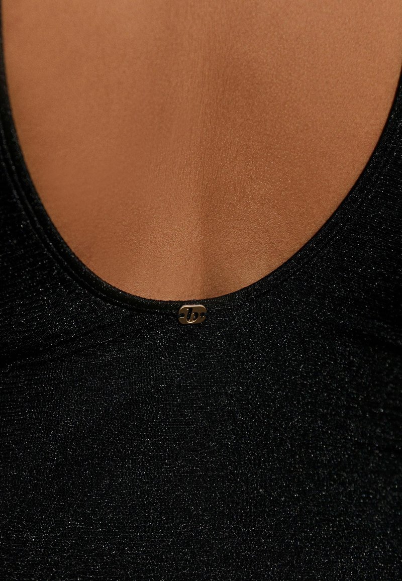 Zbliżenie na górną część klatki piersiowej osoby ubranej w czarną, teksturowaną bluzkę z okrągłym dekoltem, z małym metalicznym logo przy linii szyi.