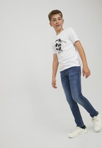 Valkoinen t-paita, jossa on jalkapallografiikka ja teksti, yhdistettynä sinisiin farkkuihin ja valkoisiin tennareihin, joissa on vihreitä yksityiskohtia. Sileä kangas, rento malli.