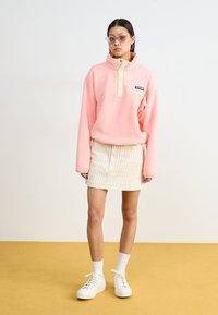 Pull en polaire rose avec col montant et patte de boutonnage beige, associé à une mini-jupe rayée beige et des baskets blanches à plateforme.
