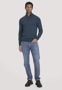 Marineblaues Zippersweatshirt aus Strickstoff, kombiniert mit hellblauen Jeans und schwarzen Schuhen. Das Modell steht aufrecht mit den Händen in den Taschen.