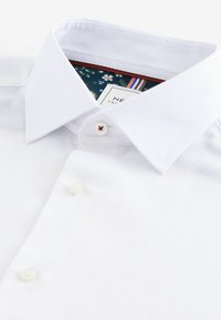 Chemise blanche avec col pointu, bouton supérieur cousu avec un fil rouge et doublure intérieure à motifs floraux visible.
