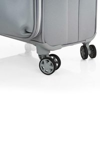 Maleta de tela blanda gris con dos ruedas negras, que presenta una superficie texturizada y un destacado detalle de cremallera blanca. Bordes redondeados y base resistente.