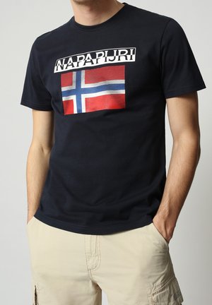 Mężczyzna w czarnej koszulce Napapijri z norweską flagą i beżowych szortach, z rękami w kieszeniach, stojący na neutralnym tle.