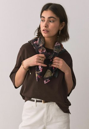Young woman wearing a dark brown top and white pants adjusts a floral scarf around her neck against a plain background.