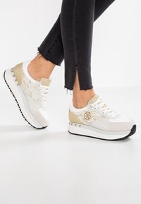 Hvide sneakers med guld accenter, snøre detaljer og en chunky sål. Har et logo på siden og dekorative nitter langs designet.